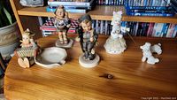 Four Hummel figurines displayed on wooden surface in front of bookshelf: a boy, two girls, and one white poodle.