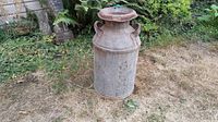 Full view of the rusty vintage metal milk can with two side handles, placed outdoors on dry grass.