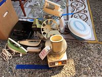 Photo showing several vintage kitchen appliances laid out including a hand mixer, small fan, iron, sandwich maker, and food processor alongside some glass bowls and Pyrex dish.