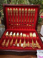 Flatware set inside a wood box with red velvet lining, showing gold-tone ornate cutting knives, soup spoons, salad forks, and dinner forks arranged in slots.