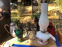 Photo showcasing white ceramic hot beverage pitcher, green vase, two glass hurricane lamps, white kerosene lamp, clear glass decorative piece, and pin all on wooden table