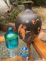 Photo showing all three items: Greek style vase, blue glass jar, and decorative glass egg on an outdoor wooden and glass surface.
