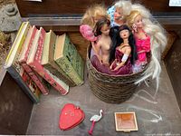 Photo shows 7 vintage children's books, 5 Barbies inside a round basket, a red heart-shaped manicure kit, white stork figurine, and a small square earring box with earrings on surface.