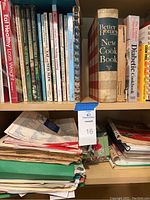 Shelf of assorted cookbooks including Better Homes and Gardens New Cook Book and health/diet cookbooks, stacked with papers beneath.