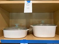 Pair of white Corningware casserole dishes, one rectangular and one square, both with clear glass lids on a shelf