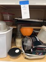 Kitchen cupboard shelf with Black & Decker food processor, orange Tupperware bowl, plastic containers, metal tray, boxed Power Steam iron, white plastic trash cans, and griddle stacked at back