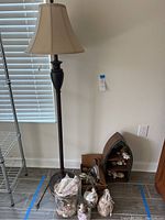 Full view of black floor lamp with cream shade next to wooden boat-shaped shelf with seashell decor and multiple glass containers filled with seashells on the floor.