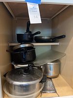 Photo showing stacked pots and pans with lids, a large cutting board, and a metal grater in a cabinet.