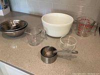 Metal, glass, and plastic measuring cups and bowls arranged on kitchen counter.