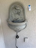 Front view of wall hanging waterfall fountain in light gray with bird and floral design, showing electric water pump and cord.