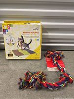 Photo showing the Kitty City Wobble Play Mat in its box and the multicolored dog rope toy lying next to it, placed on concrete background.