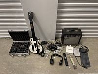 Overview of all items in the lot, including Phoenix massage gun, Guitar Hero guitar controller, video game controllers, Fender amplifier, keyboard, remote controls, headphones, Sonos device, and cables.