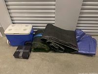 Lot showing blue Coleman cooler, rolls of black and green tarps, blue tarp, and two black plastic bins taped shut.