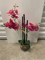 Full view of the faux orchid plant showing pink flowers, tall blackish stems, green leaves, and clear glass vase with pebbles.