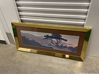 Full view of large framed landscape art print laid on floor, showing intact glass and gold frame with brown matting.