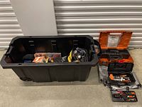 Wide shot showing large black plastic bin with assorted tools inside and four tool cases next to it on concrete floor