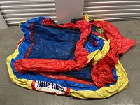 Deflated inflatable Little Tikes castle showing folded red, blue, and yellow vinyl material with visible logo laid on the floor.