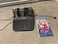 Photo of Logitech G racing pedals with three metal-faced pedals and a comic book titled 'Wire' placed on floor.