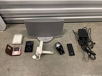 All items in the mixed electronics lot laid on a concrete floor: two iPods, Bose speaker, remote, power adapter, small microphone-shaped device, and chargers.