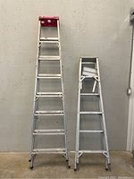 Front view of two aluminum ladders, one taller with a red top and one shorter, both showing signs of use.