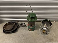 Photo showing three cast iron skillets stacked and two vintage lanterns side by side on a concrete floor.