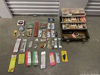 Close-up of fishing spoons and lures outside of box, showing package labels, metal finishes, and some brands.