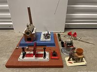 Photo shows a vintage Wilesco metal steam engine with tall chimney, flywheel, and multiple accessories including a red horn, metal blowtorch, glass container, and a pottery shaping figure.