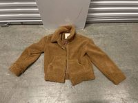 Full view of a light brown sherpa fleece jacket with collar and zipper lying flat on a concrete floor.