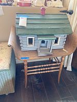Front view of the wooden log cabin dollhouse sitting on the wooden chair, showing the front porch and window details.