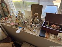 Wide shot of windowsill with assorted vintage ceramic figurines, dolls, vases, and boxes including a porcelain bust and a ceramic lidded jar.