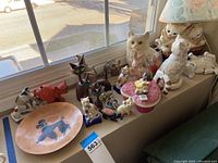 Wide view of many ceramic and wood animal figurines displayed on window ledge including cats, dogs, and others along with a cat-themed table lamp.