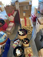 Photo showing multiple Christmas tins filled with snowman ornaments, artificial berries, holiday napkins, and assorted small decorations placed on the floor near boxes.