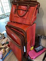 Overall photo showing two upright suitcases stacked with handbag on top, displaying red faux snakeskin material with brown handles and trim.