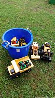 Blue plastic bucket on grass containing various toy trucks including two yellow Tonka trucks and one green John Deere tractor with damaged wheel, and other vehicles.