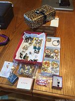 Full view of jewelry pieces, some in trays and packages, including watches, earrings, pendants, pins, small wooden box, and leopard print vinyl box