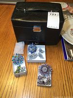 Black vinyl covered jewelry box with metal clasp and handle. Jewelry displayed in front includes several boxes containing colorful vintage costume jewelry brooches and clip-on earrings.
