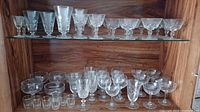 Full view of two shelves with multiple types of glassware including stemmed glasses and small glasses, all clear crystal and glass.