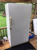Front view of a large white upright freezer placed outdoors on a deck with some miscellaneous items nearby.