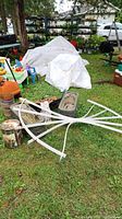 Wide view of lawn showing white double fan trellis, various garden planters, baskets, and ornaments scattered on grass near covered items and toys.