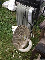 Two heaters placed on grass: a white Presto HeatDish infrared heater with gold wire grill in front and an oil-filled radiator heater behind it with power cords attached.