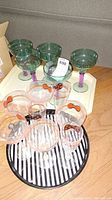 Lot showing the five green plastic martini glasses with purple stems and the white rectangular tray beneath, as well as the five clear plastic wine glasses and three champagne flutes displayed on the black and white striped round tray.