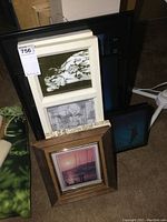 Stack of various framed prints including black, white, and wooden frames showing different artistic and photographic images.