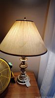Table lamp with pleated fabric shade lit, showing metal base with polished finish and decorative curves.