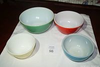 Group of 4 mixing bowls: green, red, yellow Pyrex and blue Fire King, arranged on white cloth showing top and inside views