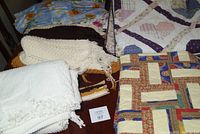 White chenille spread folded with fringed edges and a textured pattern, alongside other quilts and afghan piled on a table.