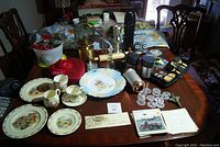 Wide view of all items arranged on a wooden table including plates, cups, a red Valentine tin box, oil lamp, cookie cutters, playing cards, and thermoses.