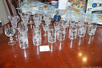 Wide view of assorted clear crystal wine glasses and tumblers arranged on a wooden table with an ornate blue glass oil lamp in the center.