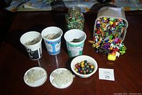 Lot displayed on a table showing glass marbles in a jar, three containers with white sea glass, small clay marbles, and assorted Kinder Egg toys in a plastic bin spilling over alongside a yellow capsule.