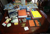 Photo showing a variety of office supplies laid out on a wooden table including calculator, notebooks, pencil case, pens, paper clips, post-it notes, and small stapler.