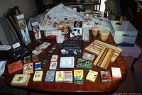 Overall view of the lot items arranged on a wooden table including boxes of wine aerator and chill stick, cutting boards, playing cards, bongo drums, watch box and medical kit.